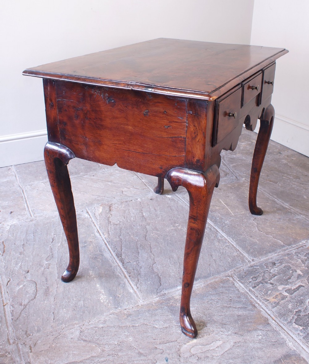 An 18th-century yew wood lowboy table featuring three drawers, a shaped scalloped apron, metal knob handles, and cabriole legs. View X