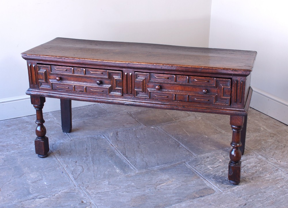 A 17th-century dark oak dresser base featuring geometric mitred mouldings across two drawers, turned front legs, and wooden drawer knobs. View IV