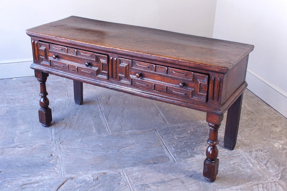 A 17th-century dark oak dresser base featuring geometric mitred mouldings across two drawers, turned front legs, and wooden drawer knobs.