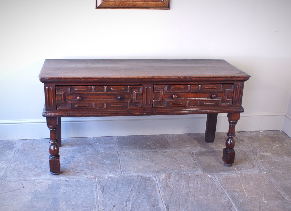 A 17th-century dark oak dresser base featuring geometric mitred mouldings across two drawers, turned front legs, and wooden drawer knobs. View V