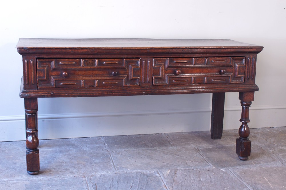 A 17th-century dark oak dresser base featuring geometric mitred mouldings across two drawers, turned front legs, and wooden drawer knobs. View XII