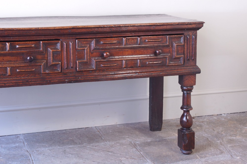 A 17th-century dark oak dresser base featuring geometric mitred mouldings across two drawers, turned front legs, and wooden drawer knobs. View XIII