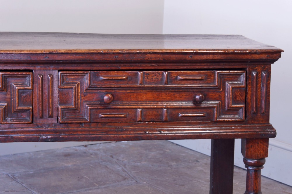 A 17th-century dark oak dresser base featuring geometric mitred mouldings across two drawers, turned front legs, and wooden drawer knobs. View XV