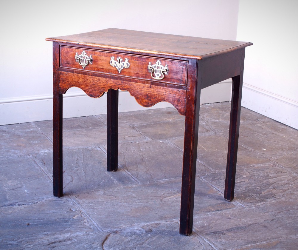 An 18th-century Georgian oak side table featuring a single drawer with decorative brass backplates and handles, set above a shaped apron and straight legs.
