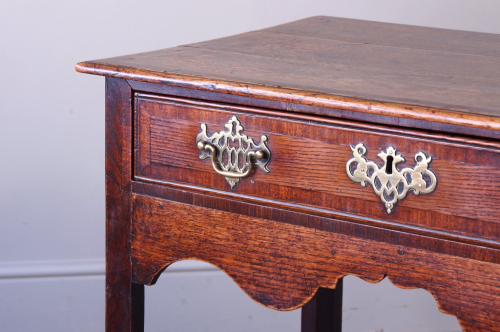 An 18th-century Georgian oak side table featuring a single drawer with decorative brass backplates and handles, set above a shaped apron and straight legs. View V