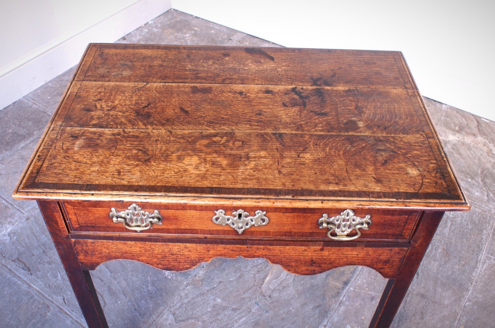 An 18th-century Georgian oak side table featuring a single drawer with decorative brass backplates and handles, set above a shaped apron and straight legs. View X