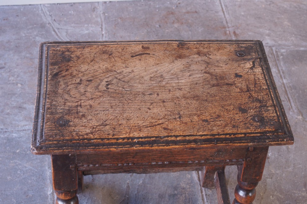 A dark oak seventeenth-century joint stool featuring turned legs, a carved frieze, perimeter stretchers, and wooden peg joinery. View III