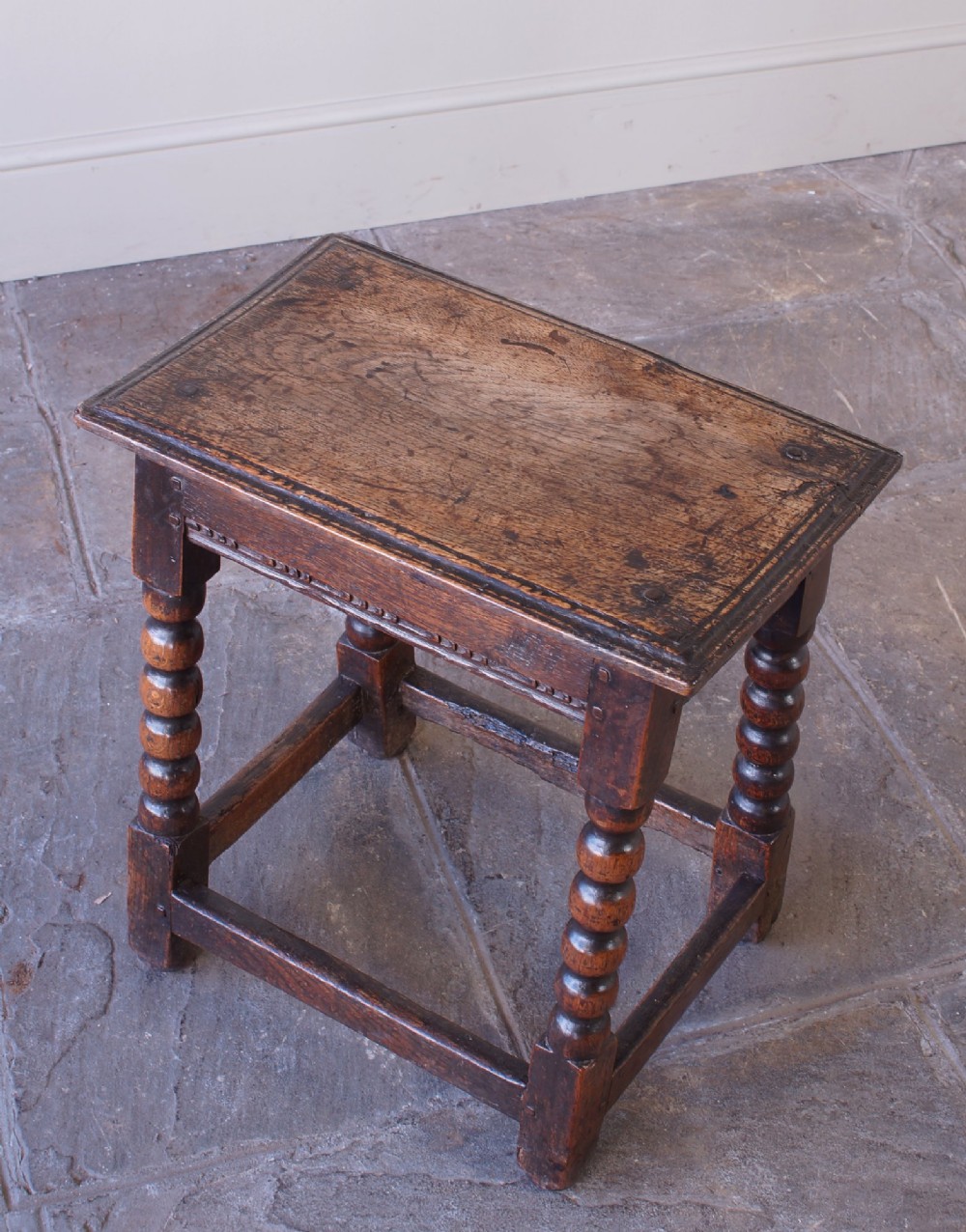 A dark oak seventeenth-century joint stool featuring turned legs, a carved frieze, perimeter stretchers, and wooden peg joinery.