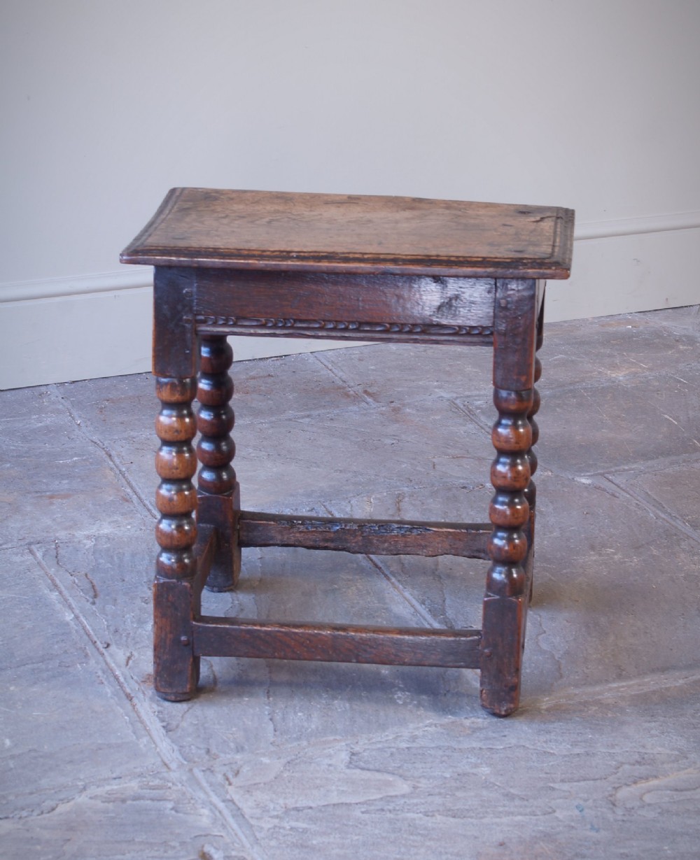 A dark oak seventeenth-century joint stool featuring turned legs, a carved frieze, perimeter stretchers, and wooden peg joinery. View IV