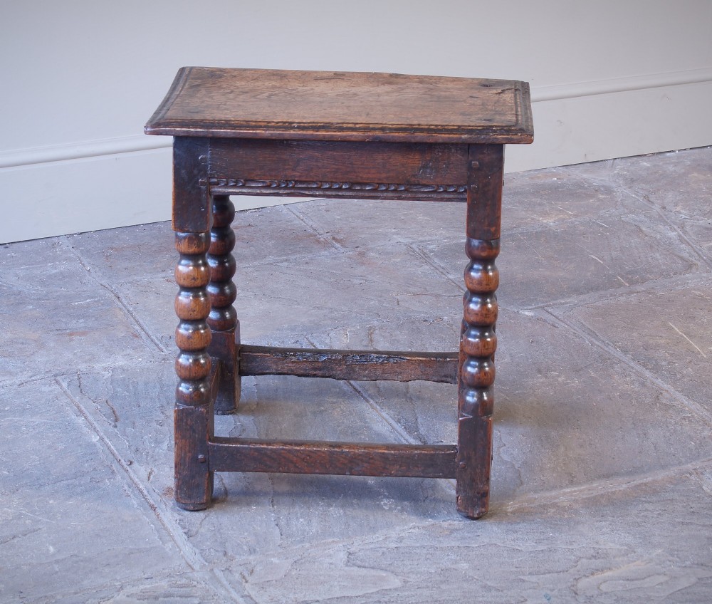 A dark oak seventeenth-century joint stool featuring turned legs, a carved frieze, perimeter stretchers, and wooden peg joinery. View V