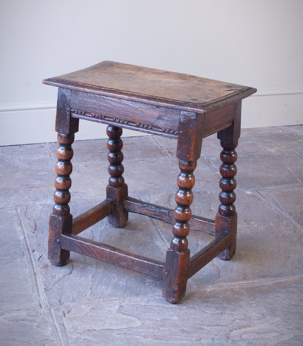 A dark oak seventeenth-century joint stool featuring turned legs, a carved frieze, perimeter stretchers, and wooden peg joinery. View II