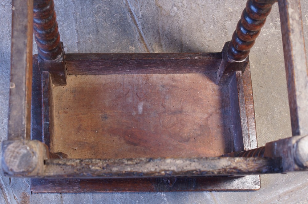 A dark oak seventeenth-century joint stool featuring turned legs, a carved frieze, perimeter stretchers, and wooden peg joinery. View VI
