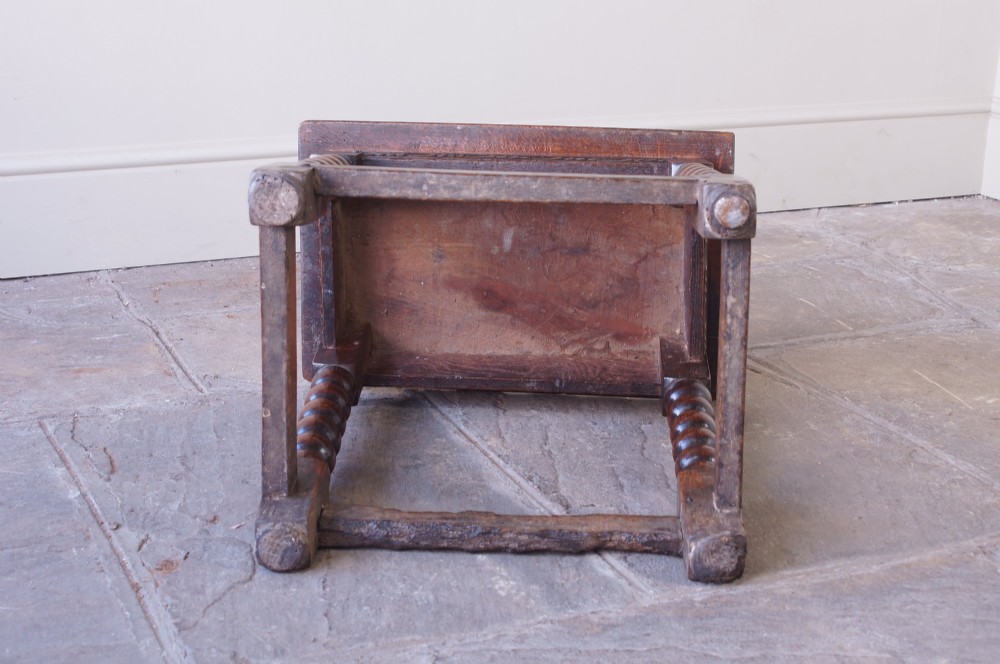 A dark oak seventeenth-century joint stool featuring turned legs, a carved frieze, perimeter stretchers, and wooden peg joinery. View VII