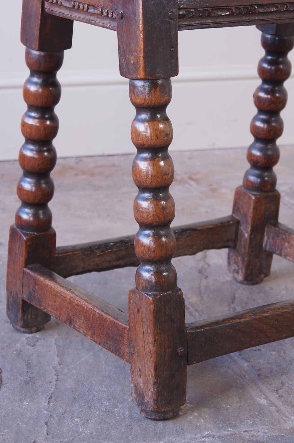 A dark oak seventeenth-century joint stool featuring turned legs, a carved frieze, perimeter stretchers, and wooden peg joinery. View VIII