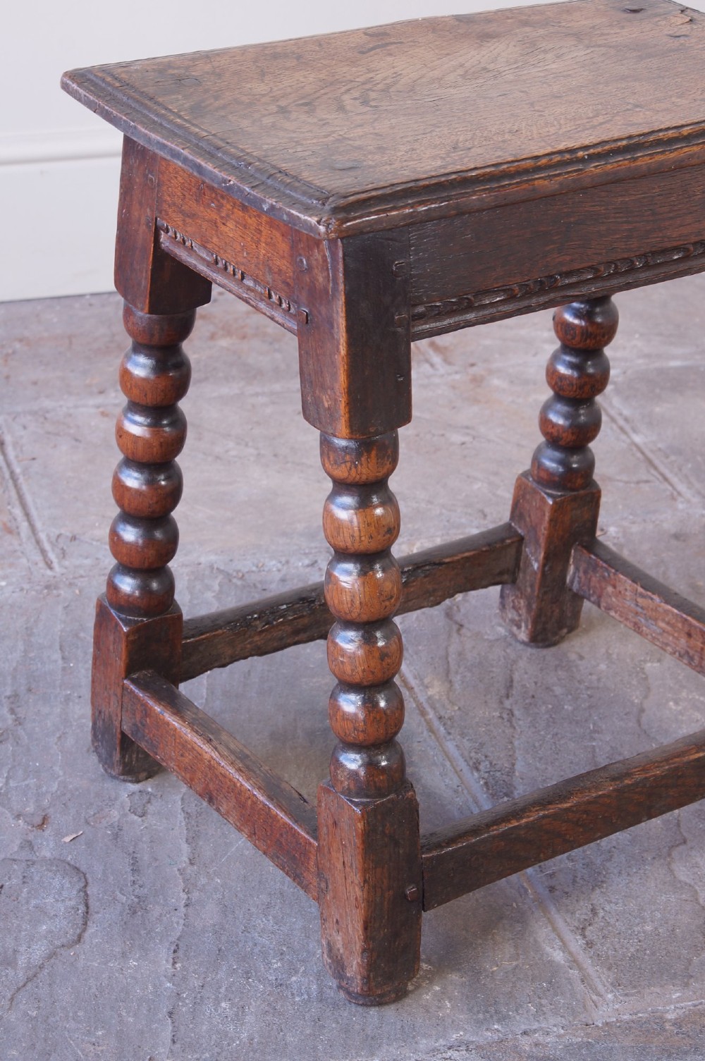 A dark oak seventeenth-century joint stool featuring turned legs, a carved frieze, perimeter stretchers, and wooden peg joinery. View IX