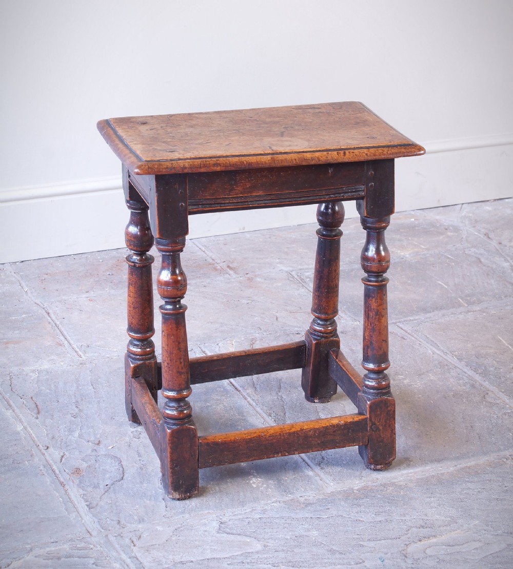 A 17th-century walnut joint stool featuring a rectangular moulded top, baluster turned legs, square pegged joinery, and a lower stretcher base with a dark patina. View II