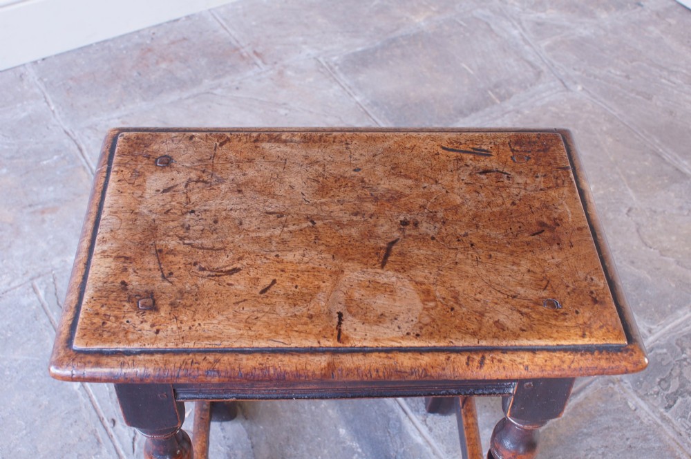 A 17th-century walnut joint stool featuring a rectangular moulded top, baluster turned legs, square pegged joinery, and a lower stretcher base with a dark patina. View III