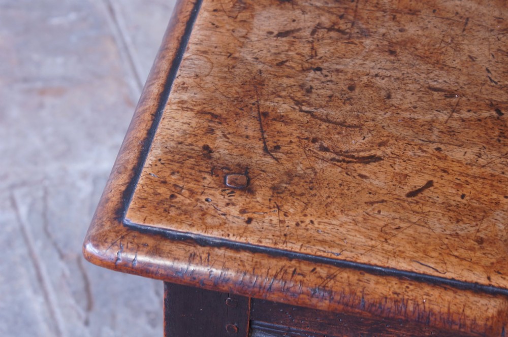 A 17th-century walnut joint stool featuring a rectangular moulded top, baluster turned legs, square pegged joinery, and a lower stretcher base with a dark patina. View VI