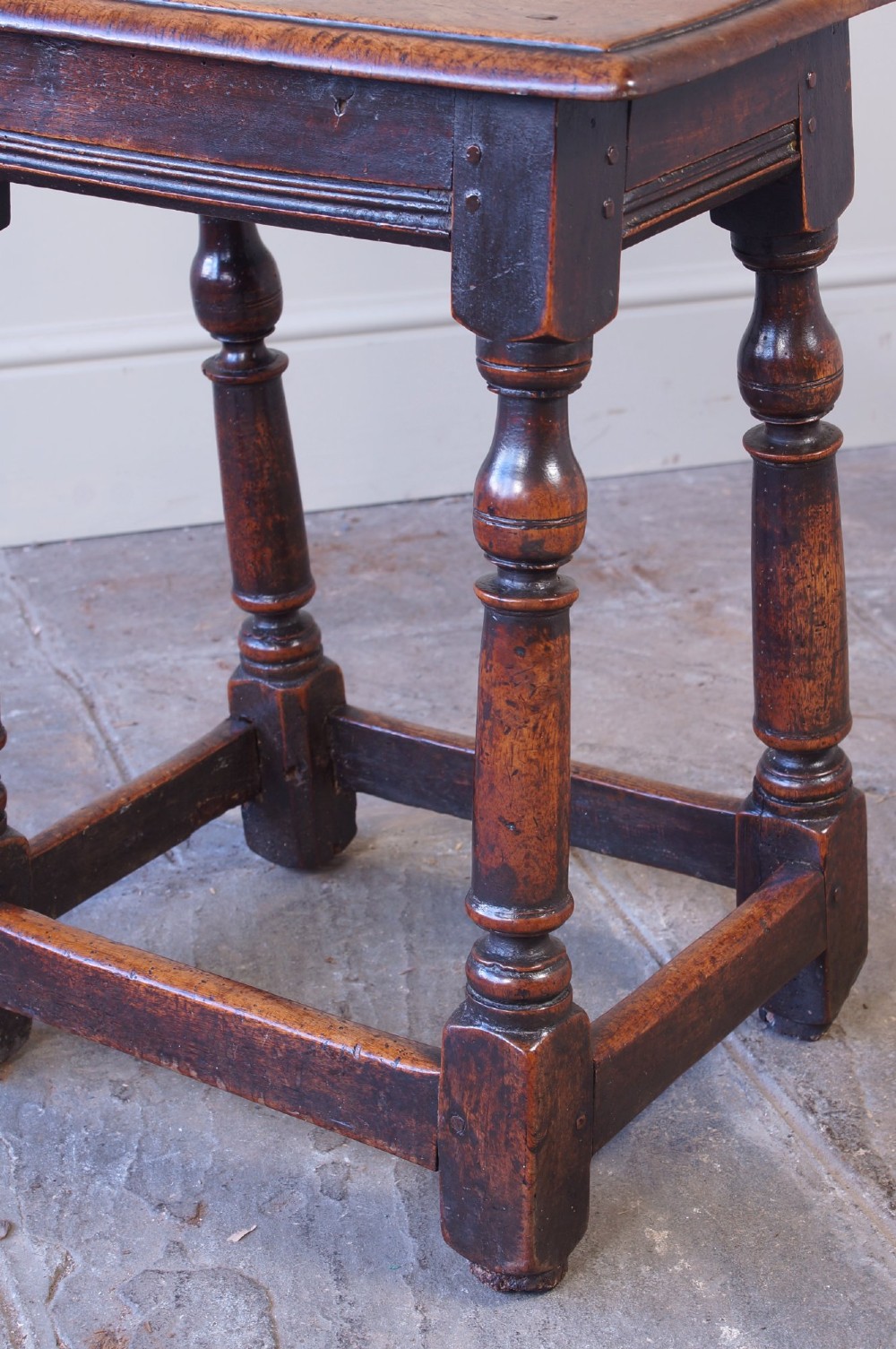 A 17th-century walnut joint stool featuring a rectangular moulded top, baluster turned legs, square pegged joinery, and a lower stretcher base with a dark patina. View VII