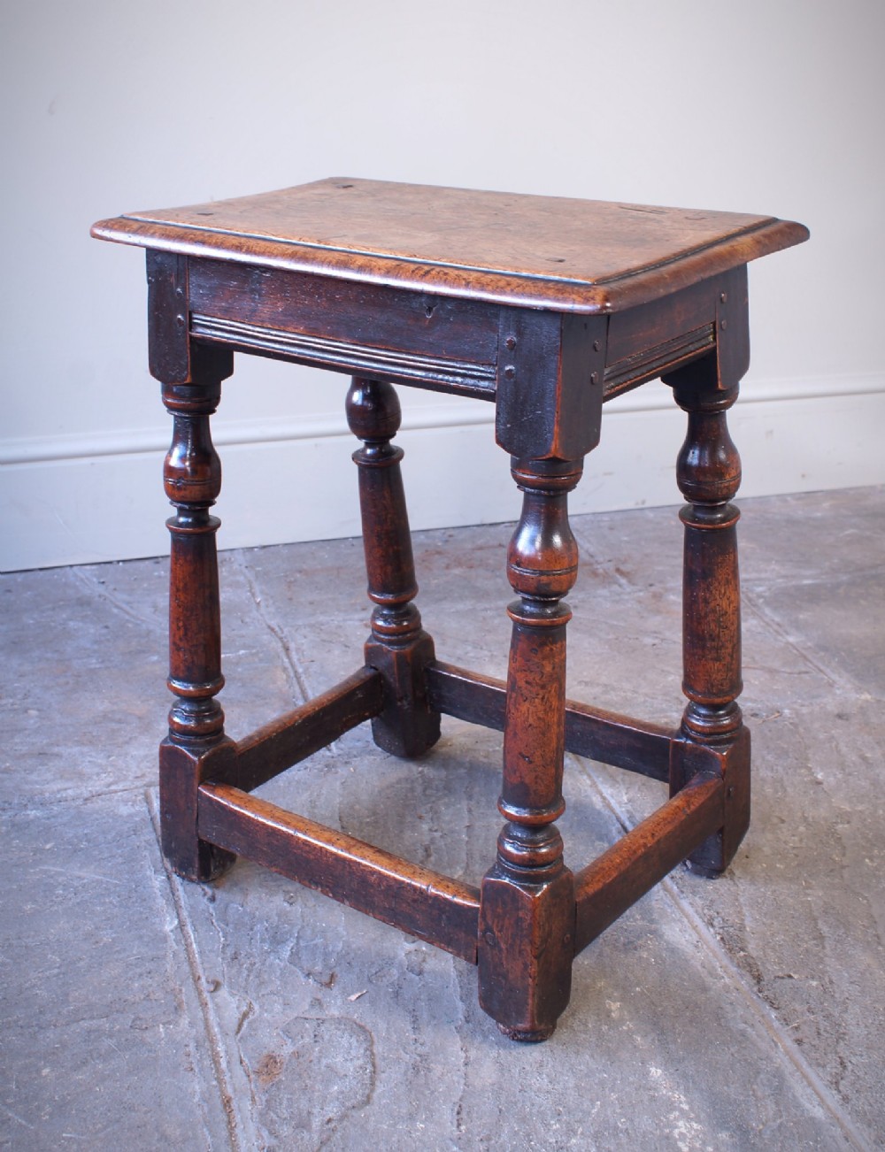A 17th-century walnut joint stool featuring a rectangular moulded top, baluster turned legs, square pegged joinery, and a lower stretcher base with a dark patina. View IX