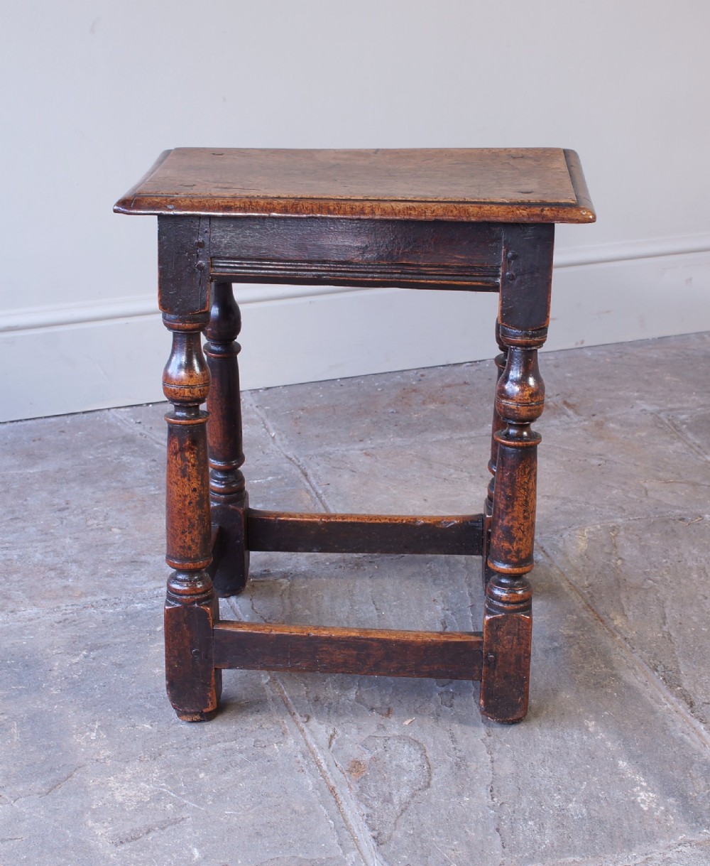 A 17th-century walnut joint stool featuring a rectangular moulded top, baluster turned legs, square pegged joinery, and a lower stretcher base with a dark patina. View X