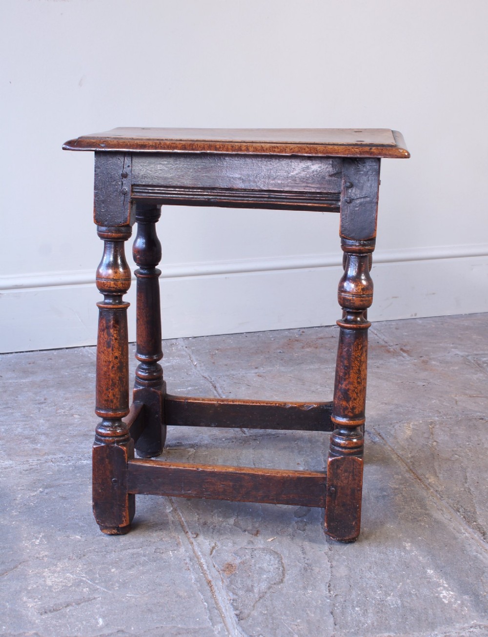 A 17th-century walnut joint stool featuring a rectangular moulded top, baluster turned legs, square pegged joinery, and a lower stretcher base with a dark patina. View XI