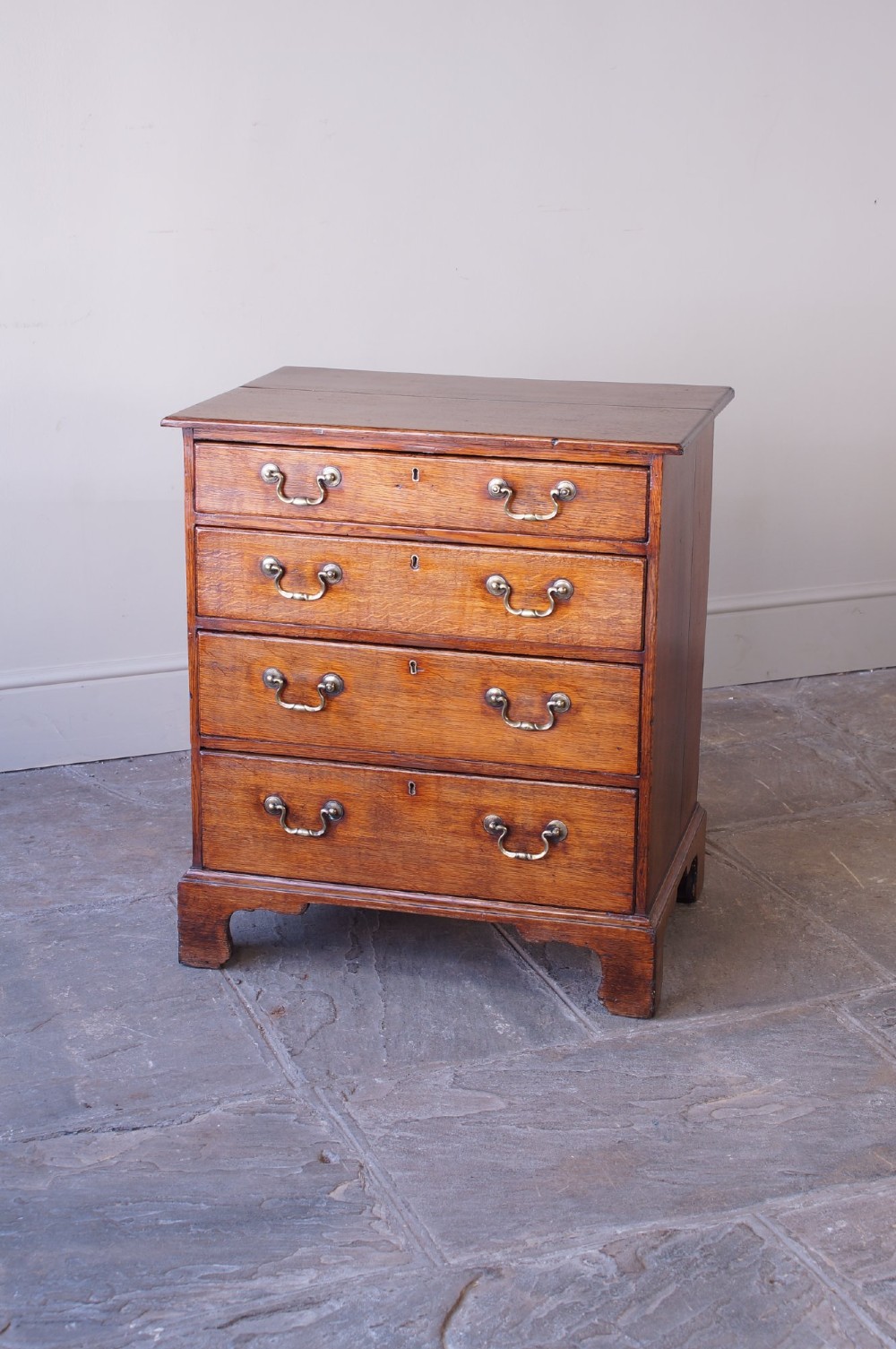 A small Georgian oak chest of drawers featuring four graduated drawers with brass swan-neck handles and bracket feet.