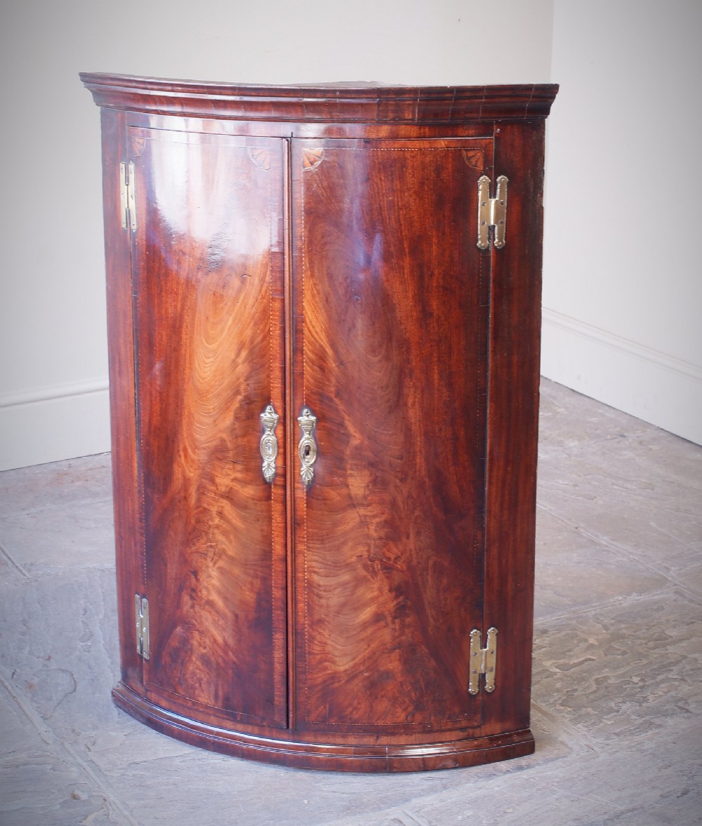 A George III bow front corner cupboard crafted from flame mahogany featuring inlaid corner spandrels, brass H-hinges, and twin curved doors.