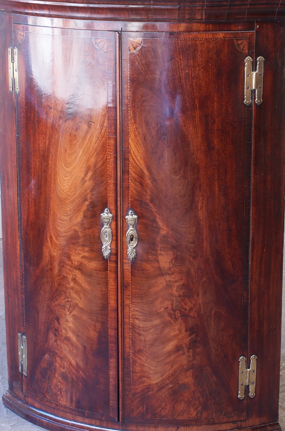 A George III bow front corner cupboard crafted from flame mahogany featuring inlaid corner spandrels, brass H-hinges, and twin curved doors. View II