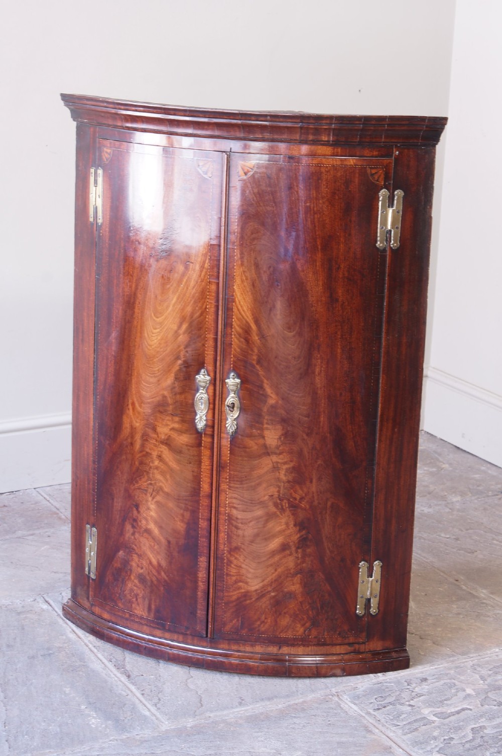 A George III bow front corner cupboard crafted from flame mahogany featuring inlaid corner spandrels, brass H-hinges, and twin curved doors. View III