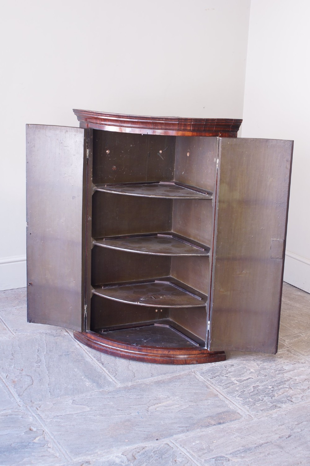 A George III bow front corner cupboard crafted from flame mahogany featuring inlaid corner spandrels, brass H-hinges, and twin curved doors. View V