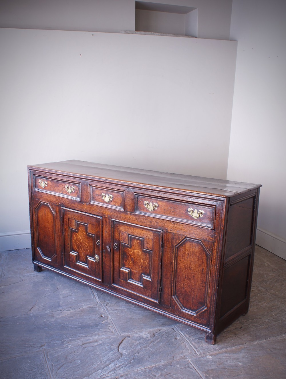 A 17th-century dark oak dresser base featuring three drawers above panelled cupboard doors with geometric moulding and brass drop handles.