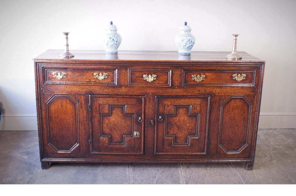 A 17th-century dark oak dresser base featuring three drawers above panelled cupboard doors with geometric moulding and brass drop handles. View II