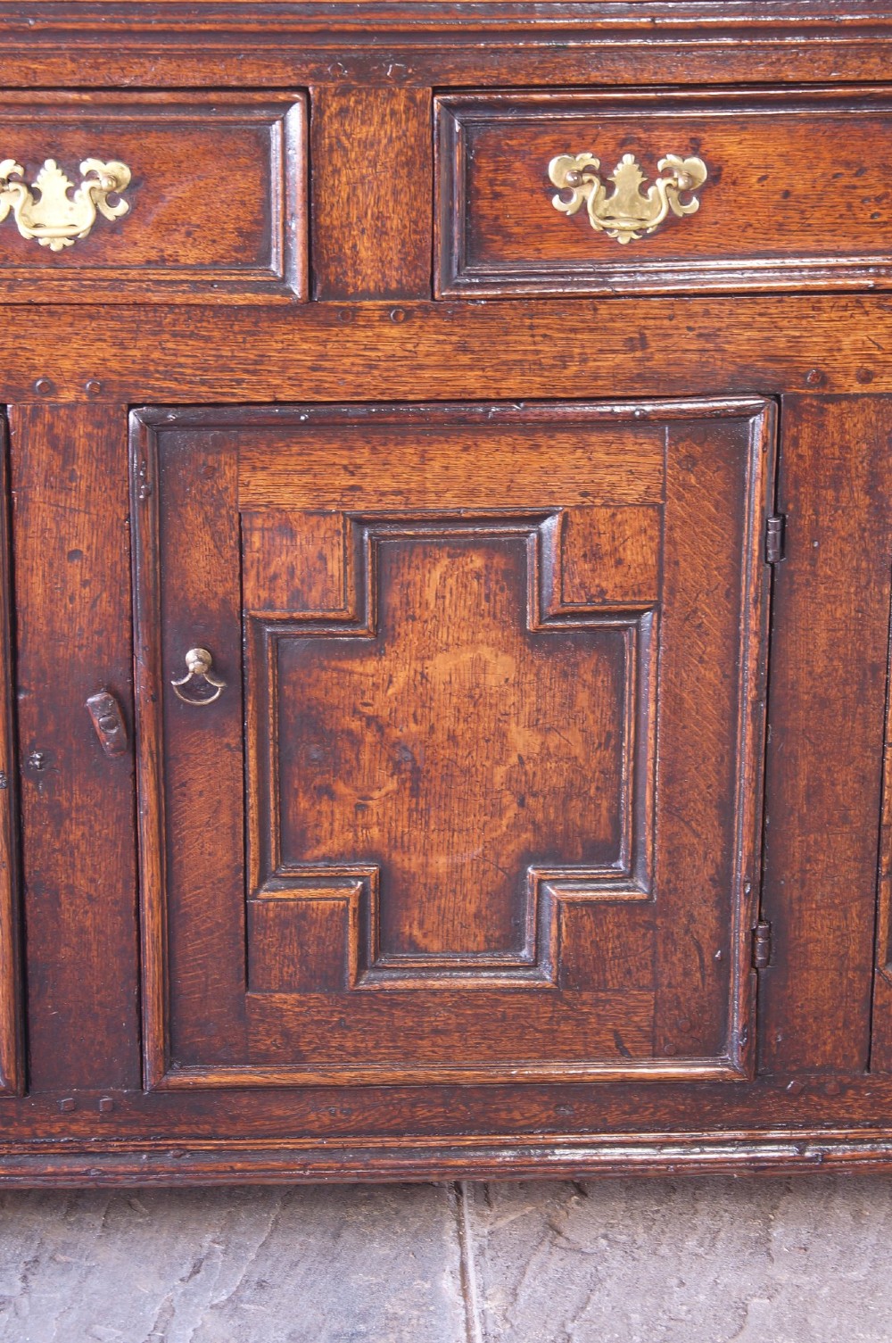 A 17th-century dark oak dresser base featuring three drawers above panelled cupboard doors with geometric moulding and brass drop handles. View III