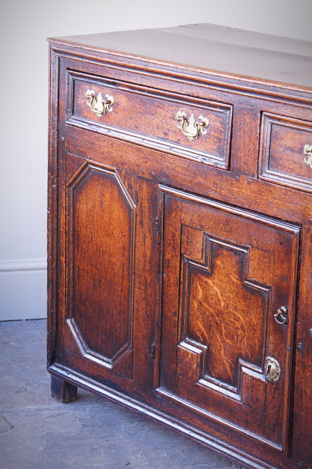 A 17th-century dark oak dresser base featuring three drawers above panelled cupboard doors with geometric moulding and brass drop handles. View IV