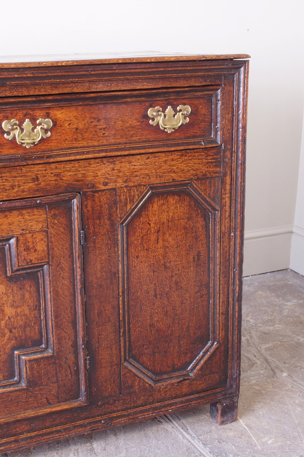 A 17th-century dark oak dresser base featuring three drawers above panelled cupboard doors with geometric moulding and brass drop handles. View V