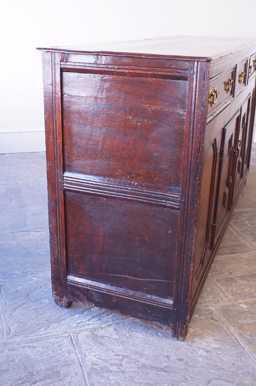 A 17th-century dark oak dresser base featuring three drawers above panelled cupboard doors with geometric moulding and brass drop handles. View VI