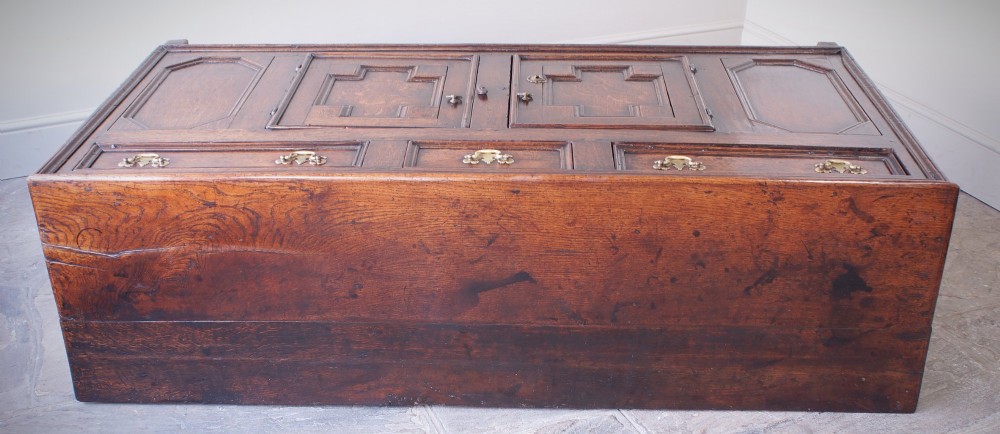 A 17th-century dark oak dresser base featuring three drawers above panelled cupboard doors with geometric moulding and brass drop handles. View VII