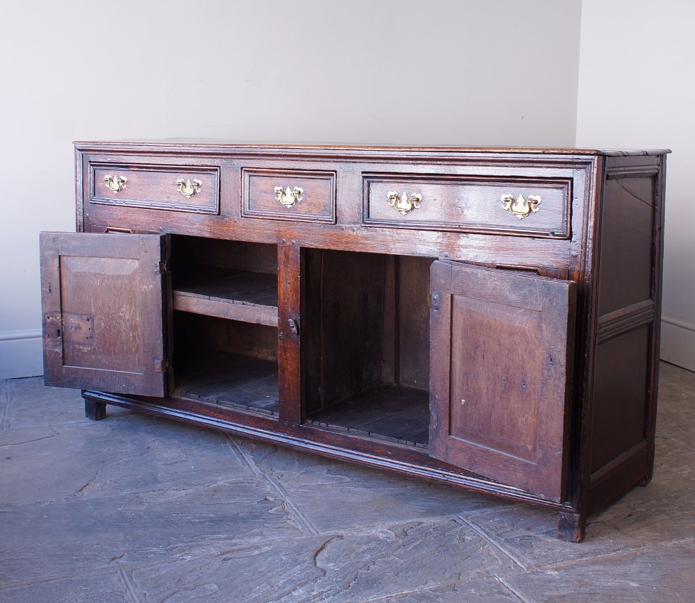 A 17th-century dark oak dresser base featuring three drawers above panelled cupboard doors with geometric moulding and brass drop handles. View X