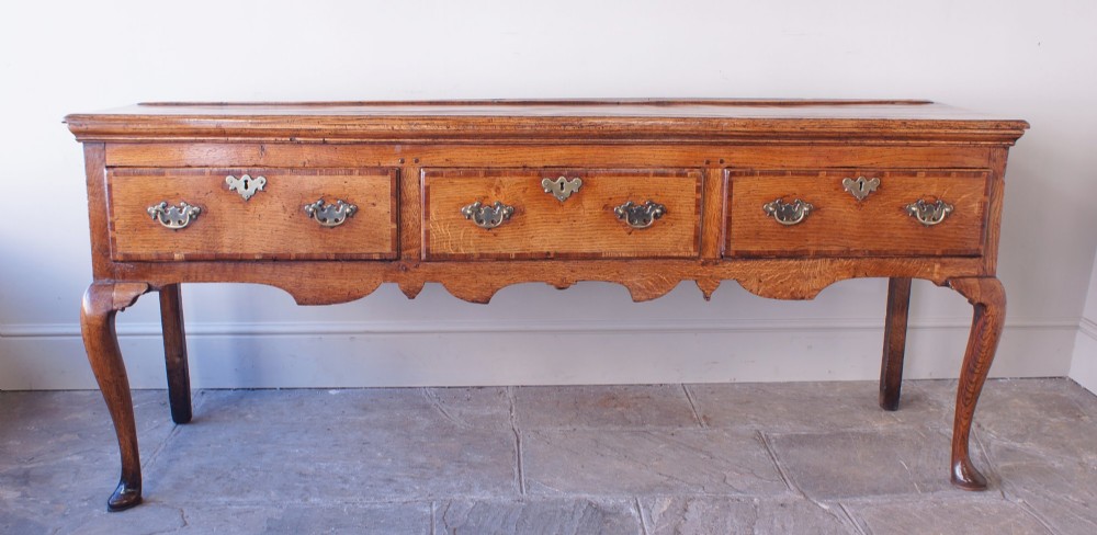 An eighteenth-century oak dresser base featuring three crossbanded drawers with brass handles, a shaped scalloped apron, and slender cabriole legs.