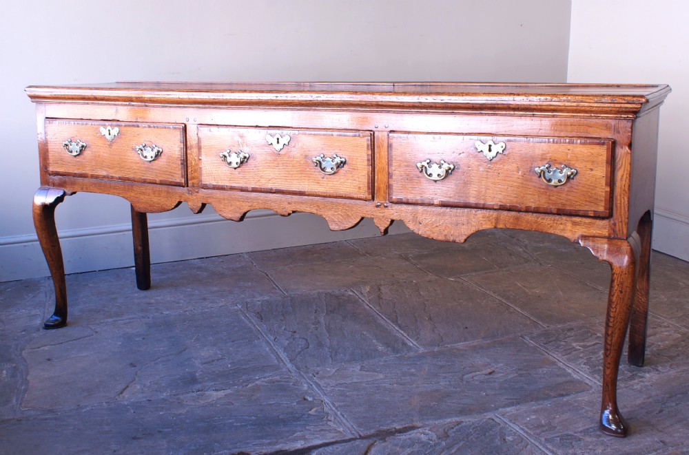 An eighteenth-century oak dresser base featuring three crossbanded drawers with brass handles, a shaped scalloped apron, and slender cabriole legs. View VI