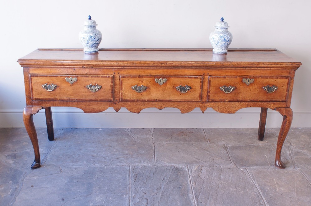 An eighteenth-century oak dresser base featuring three crossbanded drawers with brass handles, a shaped scalloped apron, and slender cabriole legs. View X