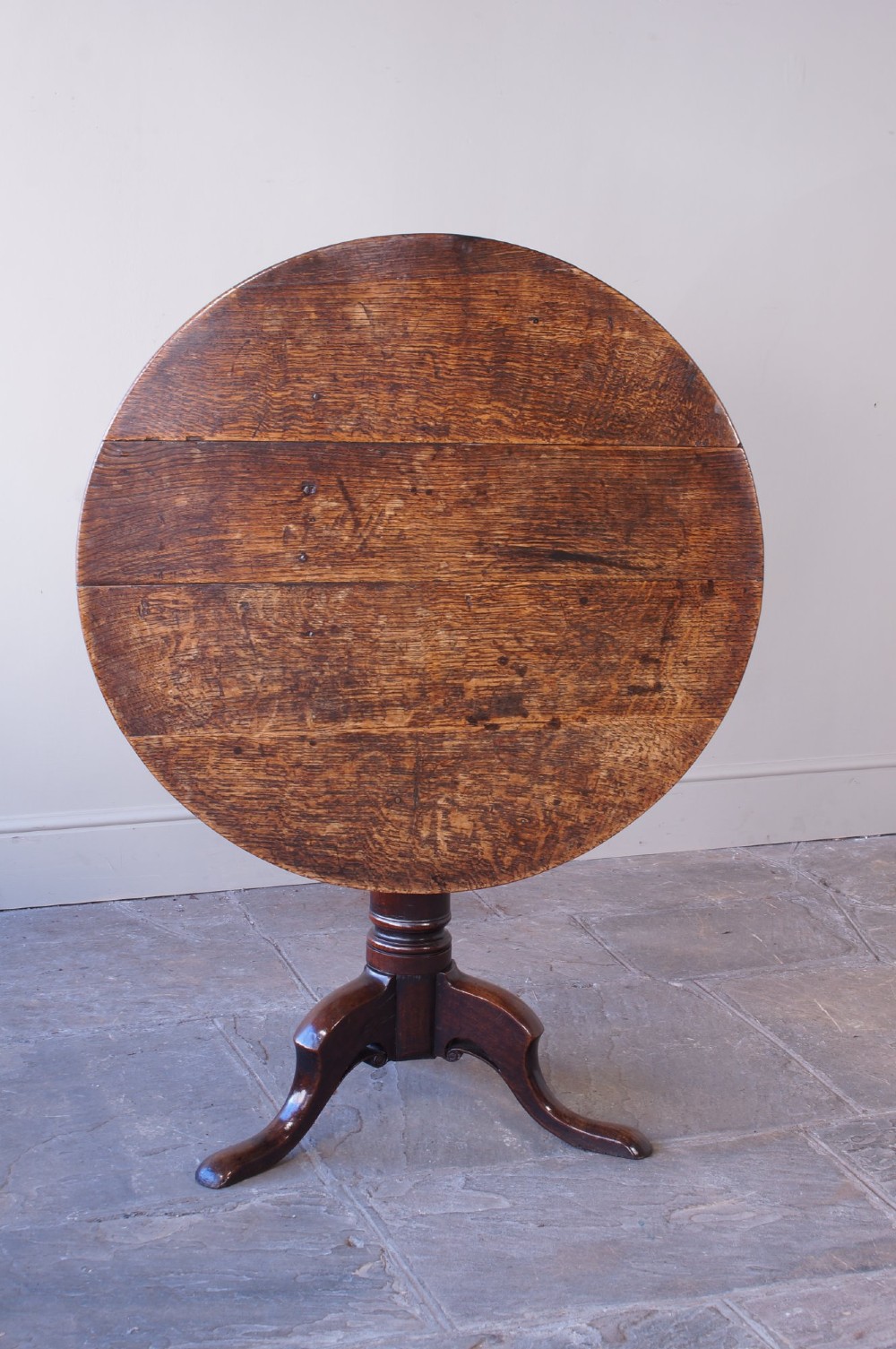 A Georgian oak tilt-top table with a circular planked top shown tilted vertically on a turned pedestal stem and tripod legs. View II