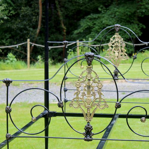 fig 1. An antique black iron four-poster canopy bed frame featuring decorative scrollwork, central brass urn medallions, and brass finials.