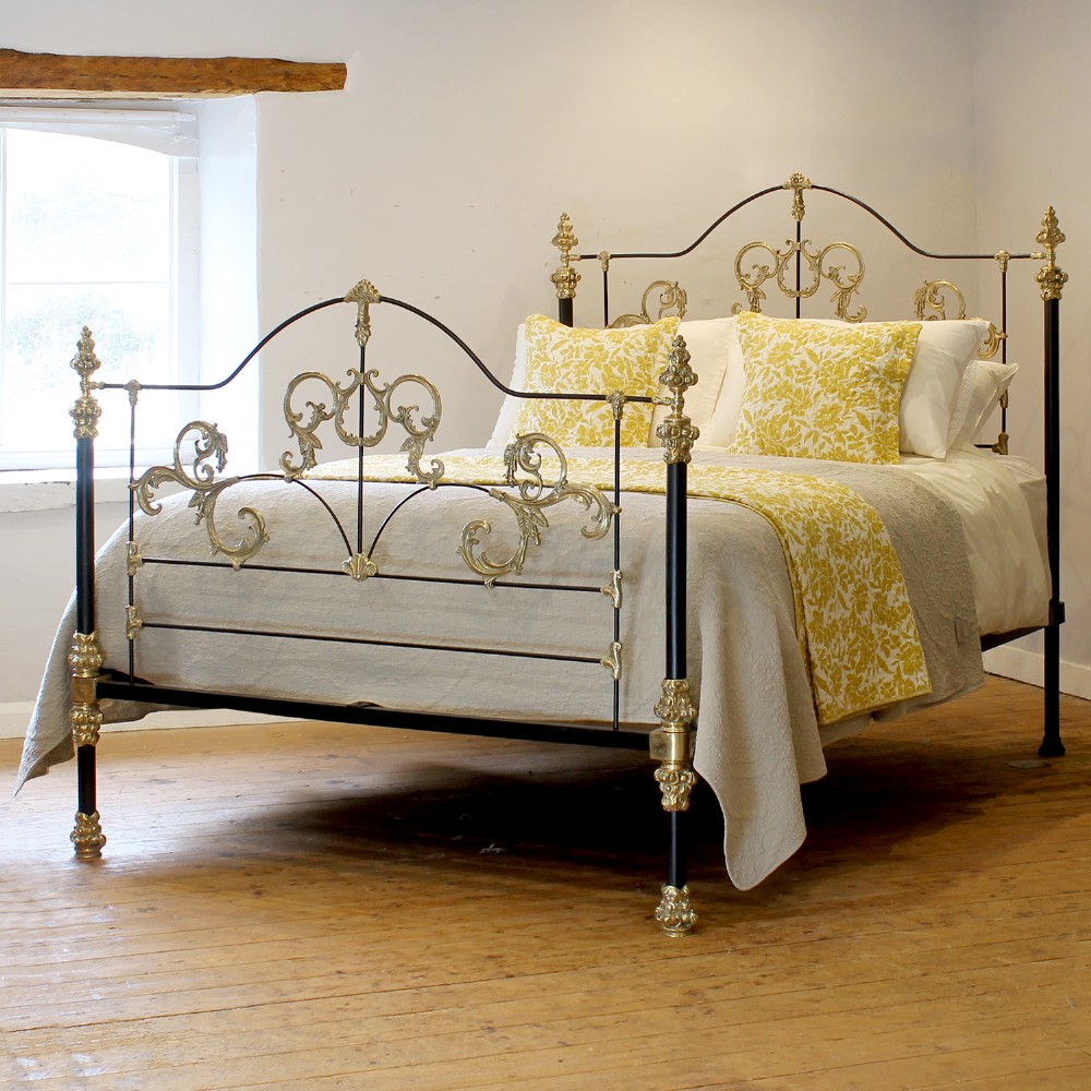 An antique king-size bedstead featuring black cast iron posts adorned with ornate scrollwork brass embellishments and decorative finials.