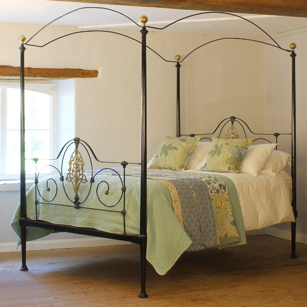 A king size Victorian black iron four poster bed featuring arched top canopy bars, decorative brass finials, and intricate cast brass scrollwork on the headboard and footboard.