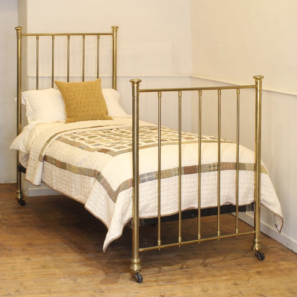 An antique Victorian single brass bedstead featuring vertical spindled headboard and footboard designs, turned finials, and frame castors.