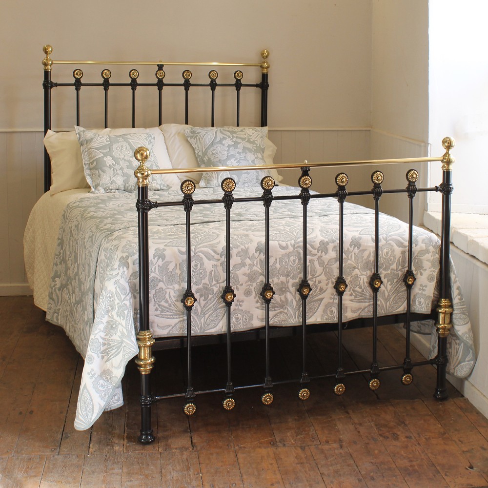 A Victorian double bed frame crafted from black painted iron with top brass rails, decorative brass finials, and brass rosettes along the vertical spindles.