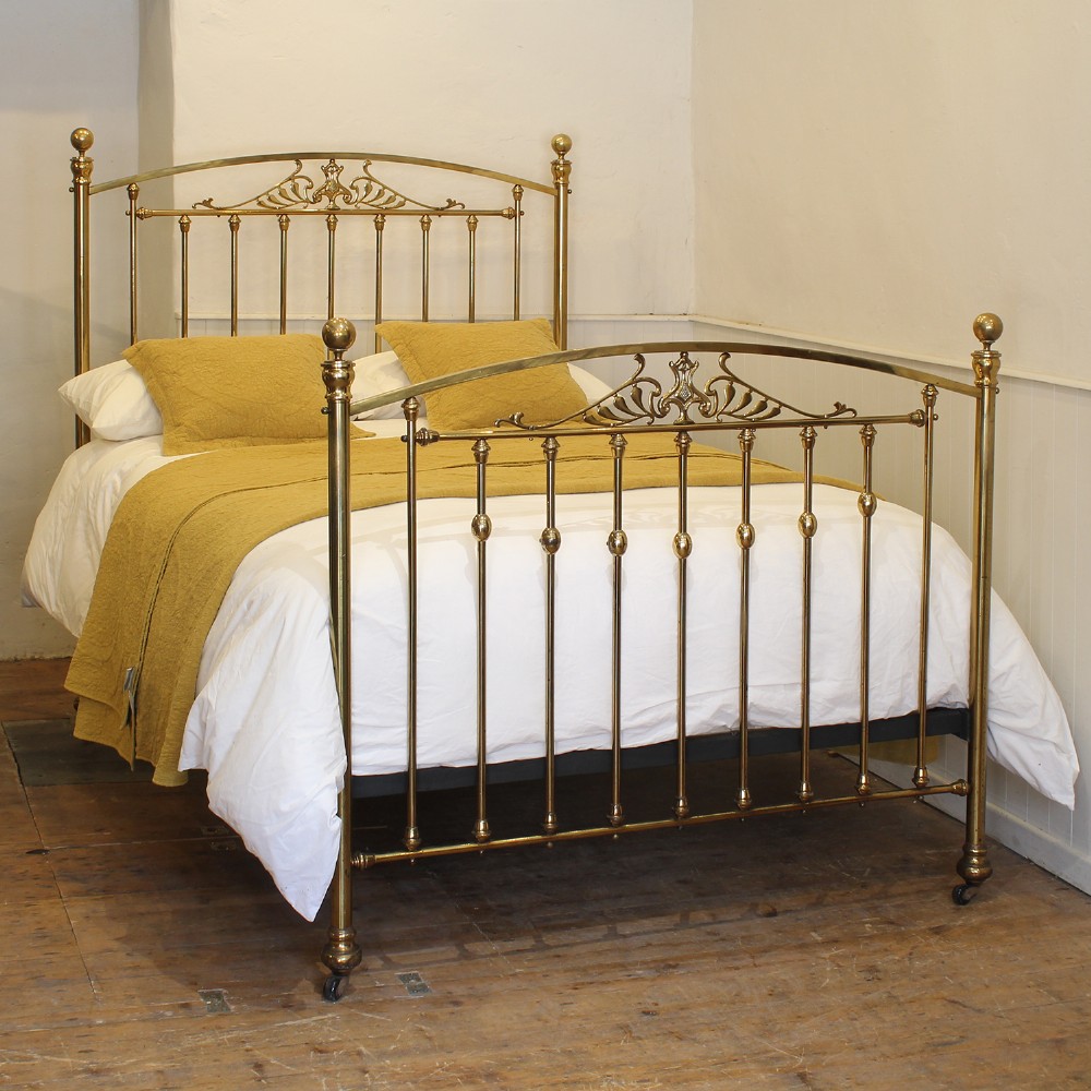 An antique double brass bed frame featuring arched headboard and footboard design with vertical spindles and central Art Nouveau scroll motifs.