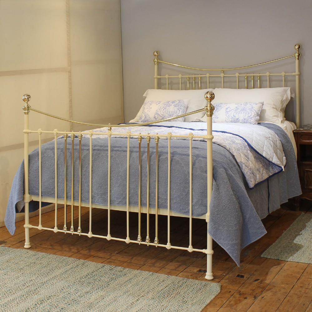 An antique king-size cream-painted metal bedstead featuring polished brass top rails, brass finials, and vertical spindle detailing.